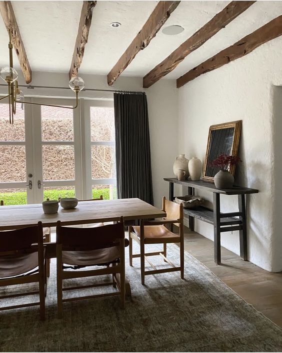 a chic earthy dining room with a glazed wall, a large stained table and leather chairs, a printed rug, a black console table and wooden beams