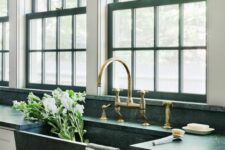 a chic farmhouse kitchen with grey cabinets and black countertops, black frame double-hung windows and gold fixtures