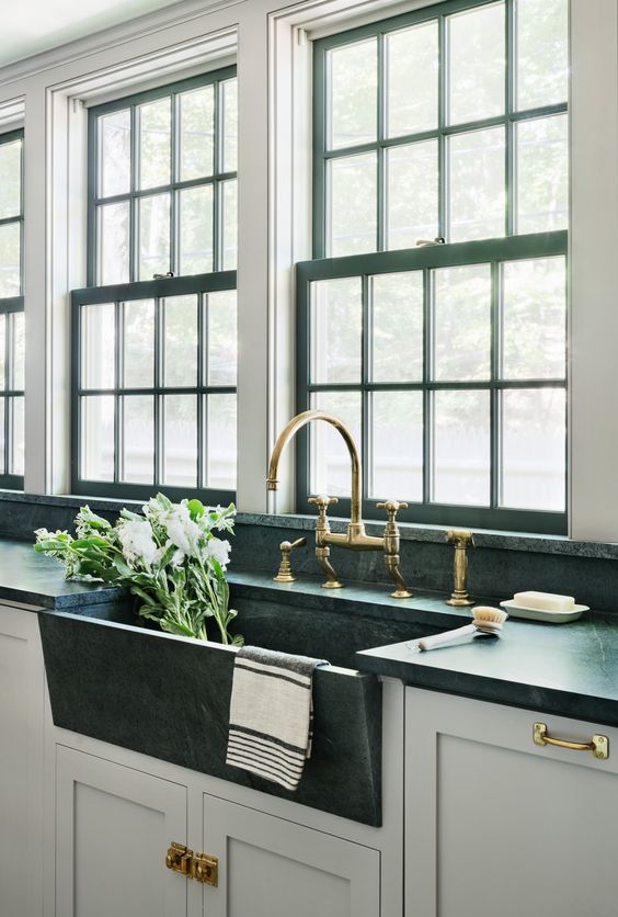 a chic farmhouse kitchen with grey cabinets and black countertops, black frame double-hung windows and gold fixtures