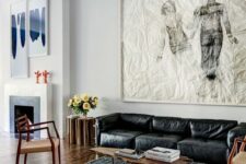 a chic living room with a black leather sofa, an oversized artwork, a black rug, a living edge table, woven chairs and a yellow lamp