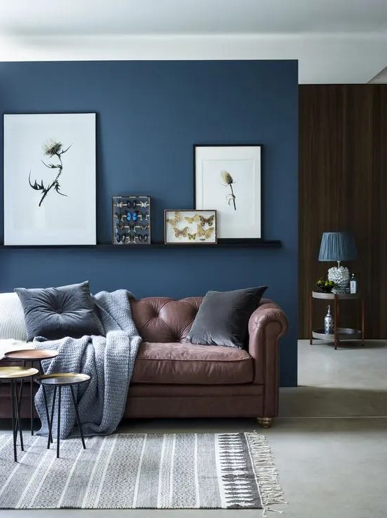 a chic living room with a brown sofa and a navy accent wall plus a ledge gallery wall and an arrangement of side tables