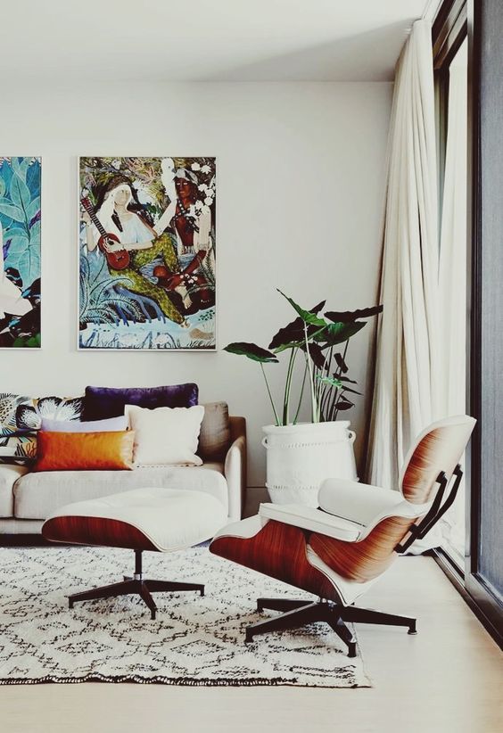 a chic living room with a neutral sofa and colored pillows, a white Eames lounger and an ottoman, a printed rug and a bright gallery wall