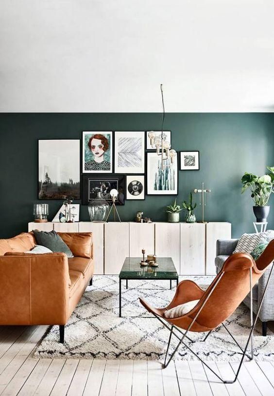 a chic mid-century modern living room with a dark green accent wall, an amber leather sofa and a butterfly chair, a coffee table and a gallery wall
