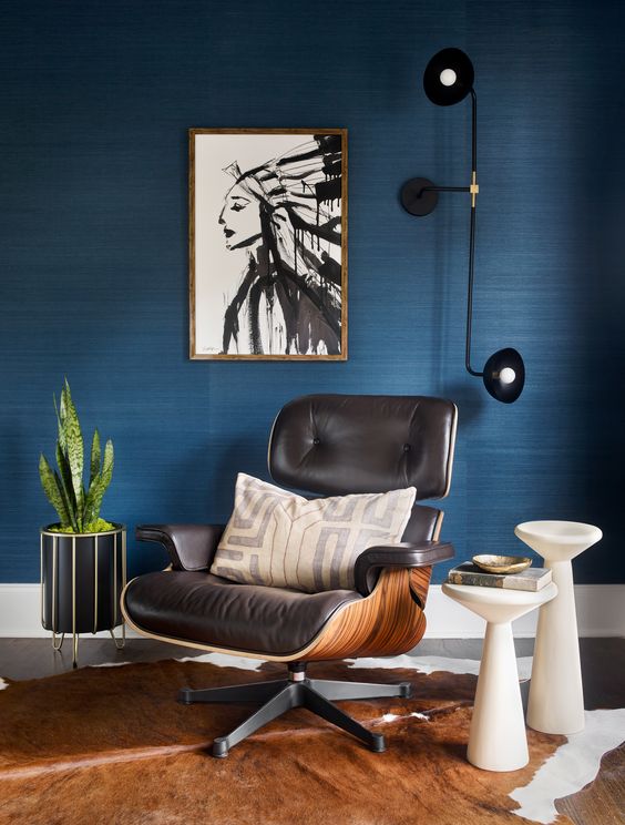 a chic mid-century modern nook with a blue grasscloth wallpaper wall, a black Eames lounger, a black sconce, white side tables and a cowhide rug