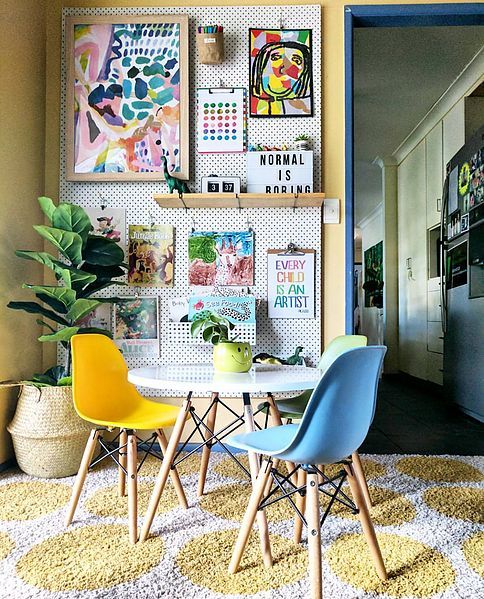 a colorful dining space with a pegboard wall with bright artwork, a round table, colorful Eames chairs, a bright printed rug