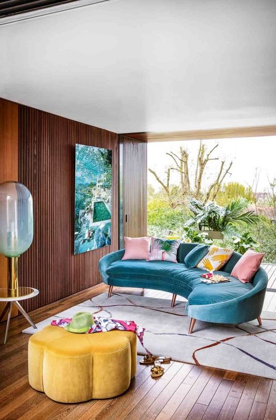 a colorful tropical living room with a bright blue velvet curved sofa, a bright yellow flower shaped ottoman and a bold artwork