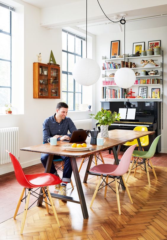 a colorful working and dining space with a trestle table, colorful Eames chairs, pendant lamps, a piano and shelves over it and a glass cabinet