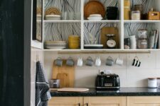 a contemporary kitchen with open white cabinets with wallpaper backing, light-stained cabinets with black countertops and large scale tiles