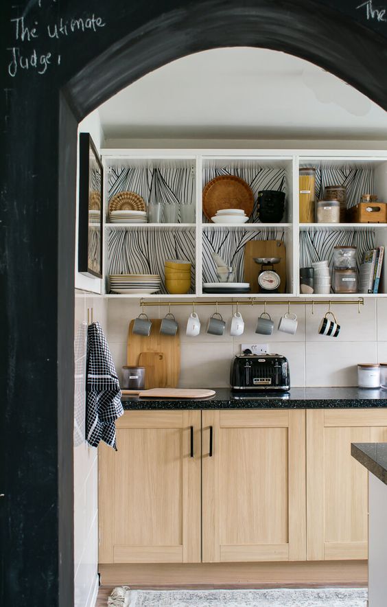 a contemporary kitchen with open white cabinets with wallpaper backing, light-stained cabinets with black countertops and large scale tiles