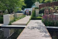a contemporary outdoor space with limed oak millboard decking, greenery and grass, a large tree and decorative ponds with pebbles
