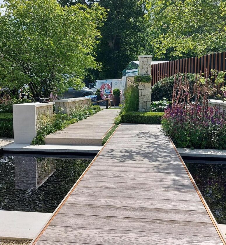 a contemporary outdoor space with limed oak millboard decking, greenery and grass, a large tree and decorative ponds with pebbles