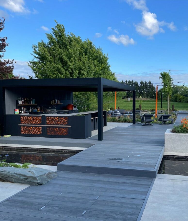 a cool modern outdoor space with brushed basalt millboard decking, a black outdoor kitchen under the roof, a pond, some greenery and rocks around