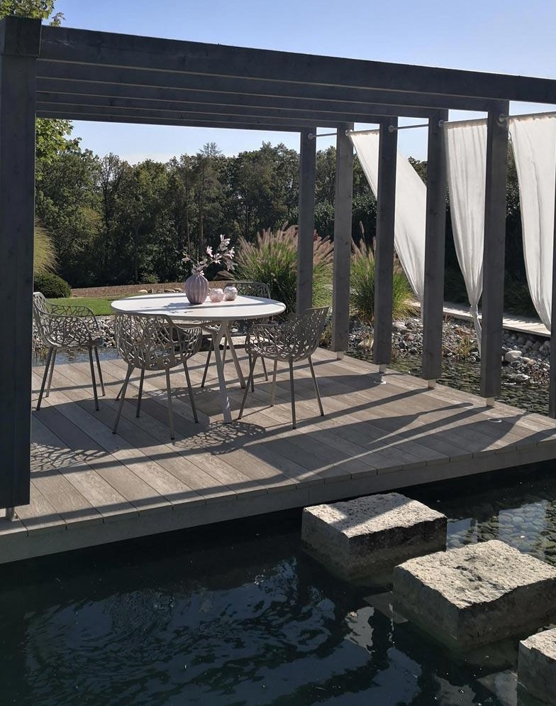 a cool terrace with a round table and woven chairs, with curtains, limed oak decking and rock steps placed right on the water