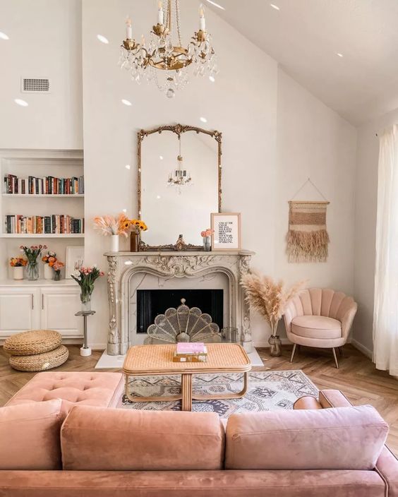 a cozy living room with a French fireplace, a rattan coffee table, a pink sectional and a chair, a gold chandelier and books