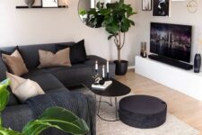 a cozy living room with grey walls, a TV unit, a black sectional and black and tan pillows, potted plants