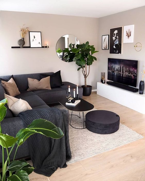 a cozy living room with grey walls, a TV unit, a black sectional and black and tan pillows, potted plants