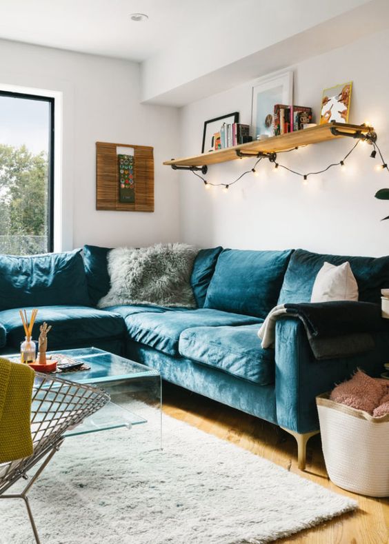 a cozy mid-century modern living room with a navy velvet sectional, an acrylic coffee table, a metal chair, a shelf with lights and some pillows