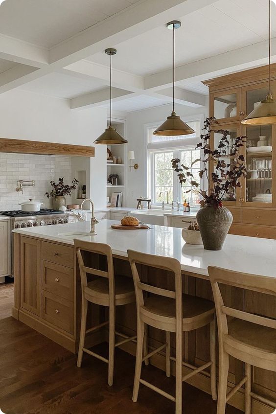 a cozy modern earthy kitchen with neutral lower cabinets, a large stained kitchen island, a large cupboard, a built-in hood and pendant lamps