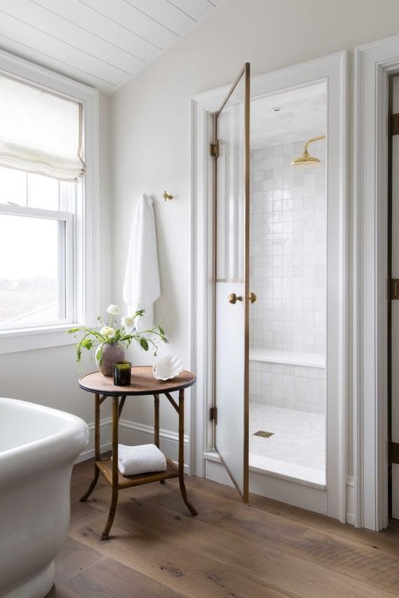 a cozy neutral farmhouse bathroom with a white tile shower space, a tub, a side table and a white double-hung window