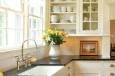 a creamy vintage kitchen with shaker and glass cabinets, black quartz countertops, a window as a backsplash and knobs and handles