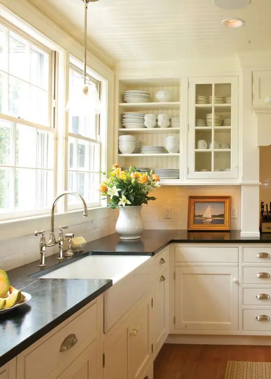 a creamy vintage kitchen with shaker and glass cabinets, black quartz countertops, a window as a backsplash and knobs and handles