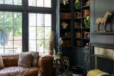a dark and moody home office with graphite grey walls and built-in bookshelves, a brown leather sofa, a navy ottoman and potted plants
