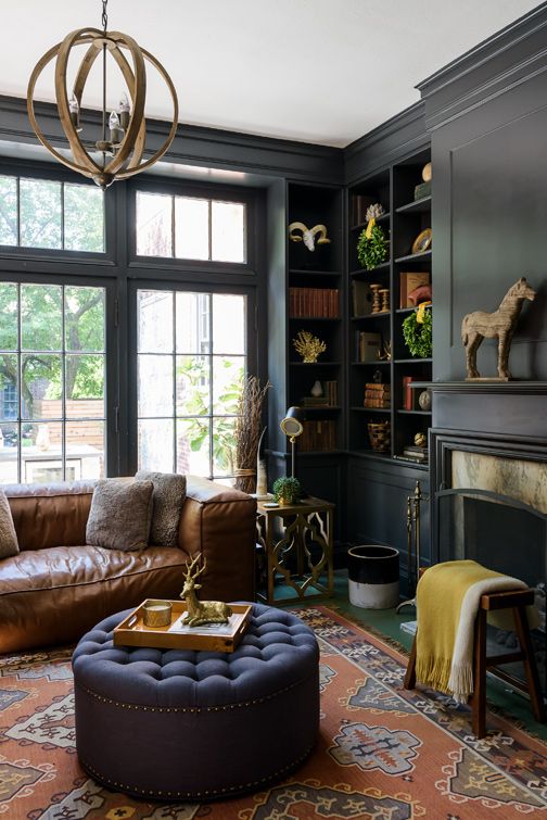 a dark and moody home office with graphite grey walls and built-in bookshelves, a brown leather sofa, a navy ottoman and potted plants