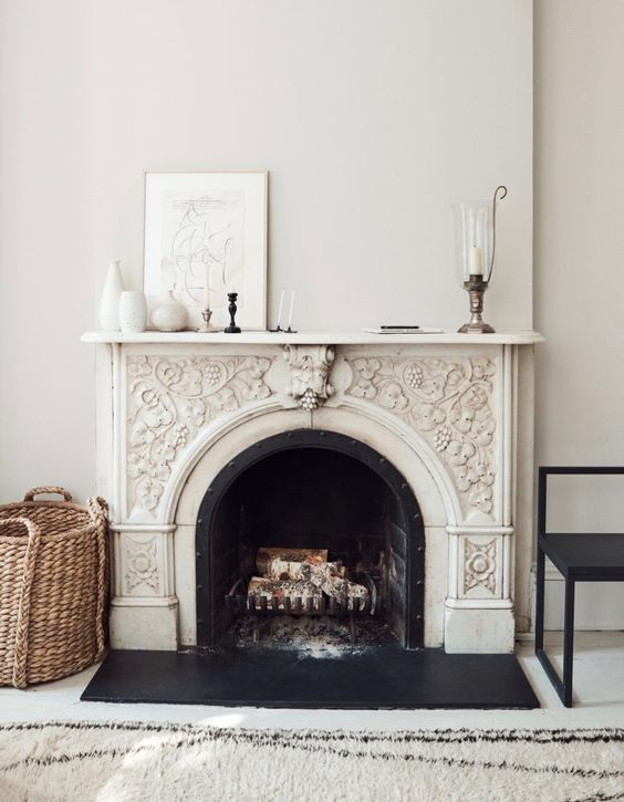 a delicately ornated French fireplace with a black chair and a basket next to it, some decor on the mantel and a printed rug