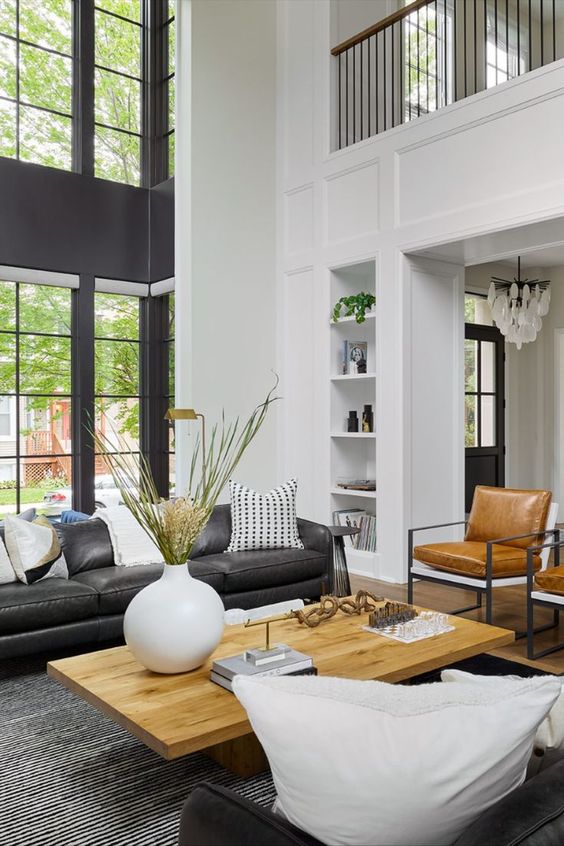 a double height living room with a black leather sofa, amber chairs, a coffee table and some built in shelves and greenery