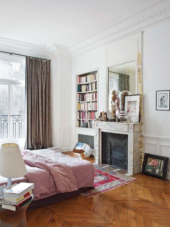 a dreamy Parisian bedroom with a French  fireplace, a bed with pink bedding, a glass nightstand, a built-in bookcase and lovely art