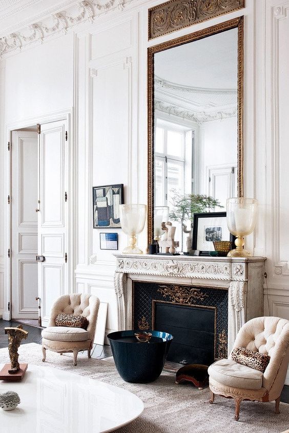 a fab neutral living room with a French fireplace, neutral chairs, an oversized mirror in a chic frame and some decor on the mantel