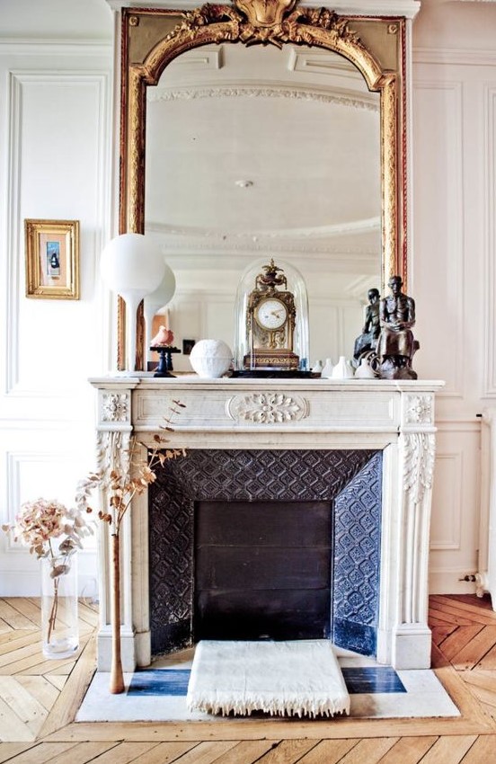 a fabulous French fireplace with an ornated white mantel, an oversized mirror and some vintage decor on the mantel
