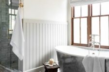 a farmhouse bathroom with paneling and penny tiles on the floor, a metal clad tub, stained double-hung windows, white towels