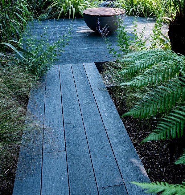 a garden with brushed basalt millboard decking, greenery and grass around and a bowl with water and a wooden lid
