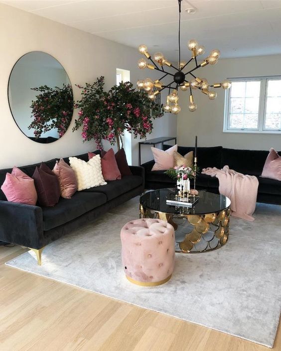 a glam living room with black sofas, pink and mauve pillows, a gold bulb chandelier, a black and gold coffee table and a pink pouf