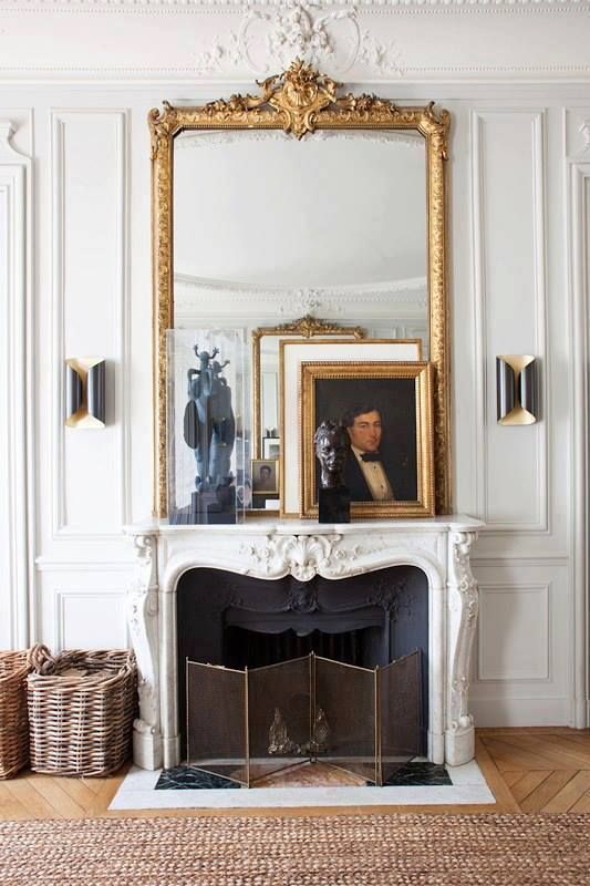 a gorgeous French fireplace with an oversized mirror, some artwork, a folding screen, baskets with firewood