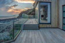 a gorgeous coastal deck done with smoked oak boards, with a cool sea view and a glass banister looks very modern and chic