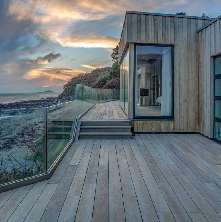 a gorgeous coastal deck done with smoked oak boards, with a cool sea view and a glass banister looks very modern and chic
