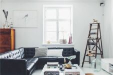 a light-filled Scandinavian living room with a black leather sectional, a low industrial coffee table, a white TV unit, a ladder as a storage piece, a rust pouf