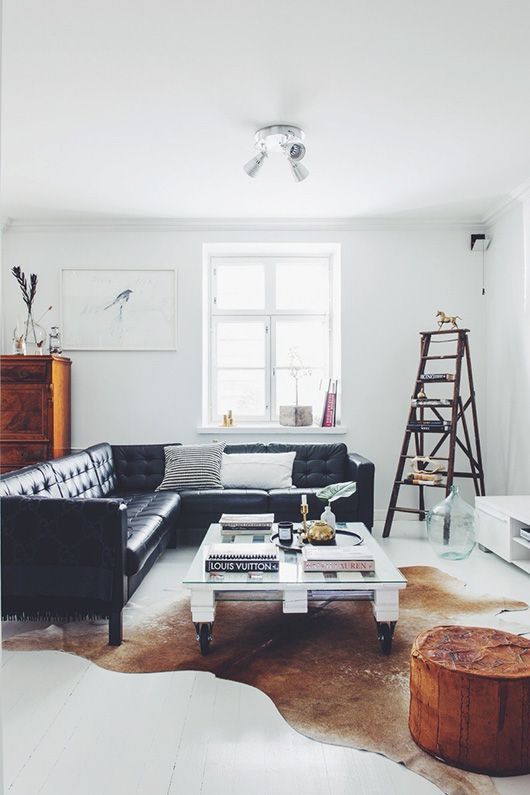 a light filled Scandinavian living room with a black leather sectional, a low industrial coffee table, a white TV unit, a ladder as a storage piece, a rust pouf