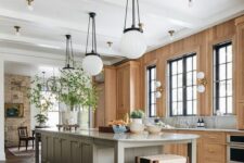 a light-filled earthy kitchen with a stained wall and cabinets, a grey kitchen island, pendant lamps and potted greenery