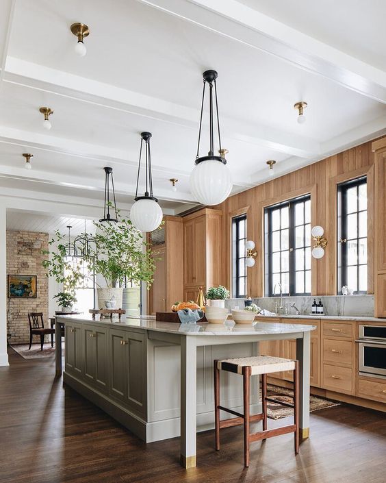 a light filled earthy kitchen with a stained wall and cabinets, a grey kitchen island, pendant lamps and potted greenery