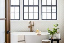 a lovely bathroom with black frame double-hung windows, an oval tub, a side table and a tree stump plus a printed rug