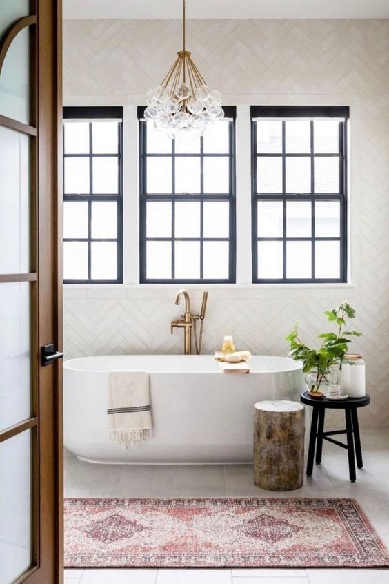 a lovely bathroom with black frame double-hung windows, an oval tub, a side table and a tree stump plus a printed rug