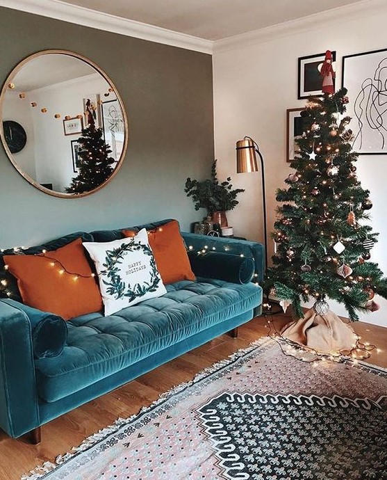 a lovely living room with a turquoise velvet sofa, a Christmas tree with lights, a floor lamp and a round mirror plus a printed rug