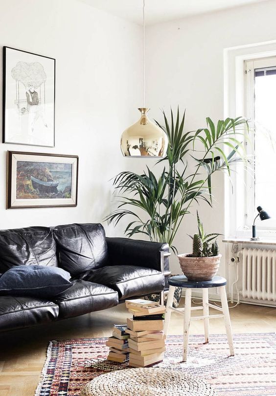 a lovely modern living room with a black leather couch, a boho printed rug, a stool, a potted plant, a copper pendant lamp and book stacks