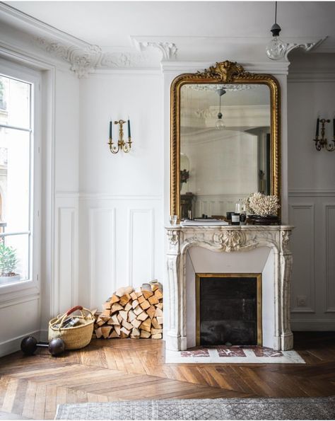 a lovely space with an ornated French fireplace, an oversized mirror in a gilded frame, firewood and a basket