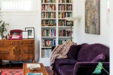 a mid-century modern living room done in neutrals and filled with color – a printed rug, a deep purple sofa, stained furniture and a built-in bookcase