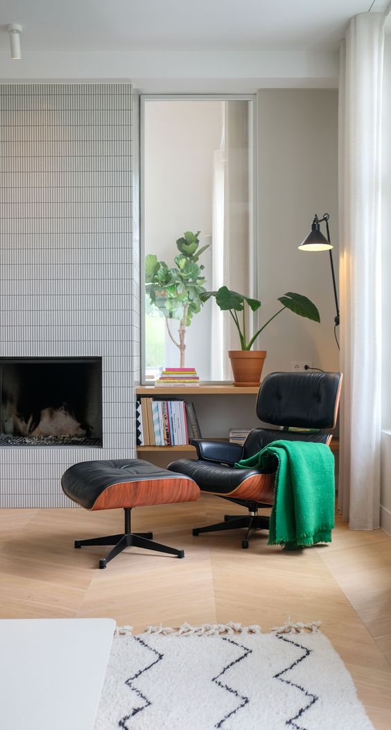 a mid-century modern nook with a fireplace clad with skinny tiles, a black Eames chair with an ottoman, built-in bookshelves