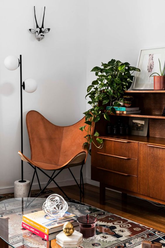a mid-century modern space with a stained sideboard, an amber leather butterfly chair, a printed rug, a glass coffee table and a plant